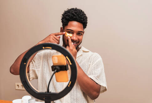 A smiling young latin man applying face patch while making a playful gesture, with a ring light and smartphone setup in view. - ADSF50013