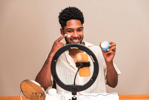 Smiling young latin man demonstrating a skincare product in front of a ring light, creating content for his beauty vlog. - ADSF50015