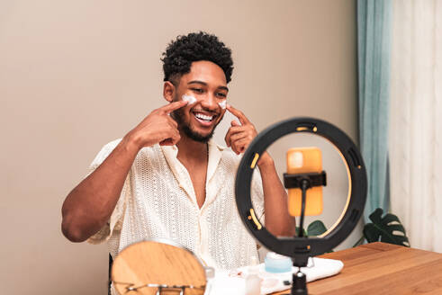 Cheerful latin man enjoying his skincare routine, smiling and pointing at cheeks in front of a ring mirror at home. - ADSF50016