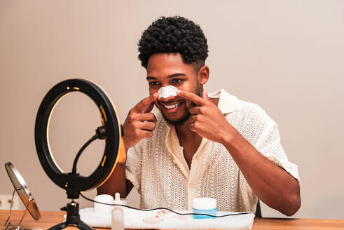 A cheerful young latin man applies a nose strip for skincare in front of a ring light, promoting self-care. - ADSF50017