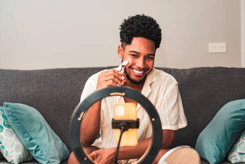 A cheerful young latin man records a skin care theme video blog on his smartphone secured to a ring light, smiling while using a facial roller on a couch at home. - ADSF50018