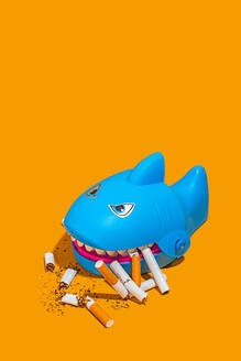 Hoher Winkel des blauen Haifischspielzeugs inmitten von verstreuten Zigarettenkippen auf gelbem Hintergrund - ADSF50023
