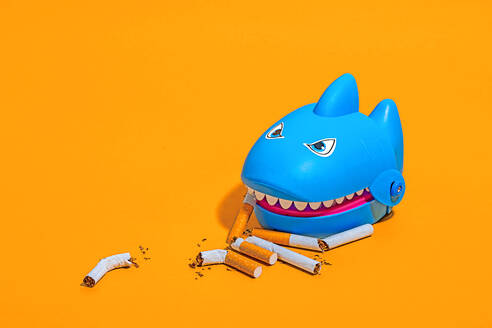 Hoher Winkel des blauen Haifischspielzeugs inmitten von verstreuten Zigarettenkippen auf gelbem Hintergrund - ADSF50024