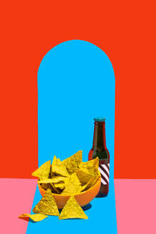 A vibrant still life image featuring a bowl of crunchy nacho chips beside a dark bottle against a split background of red and blue - ADSF50025
