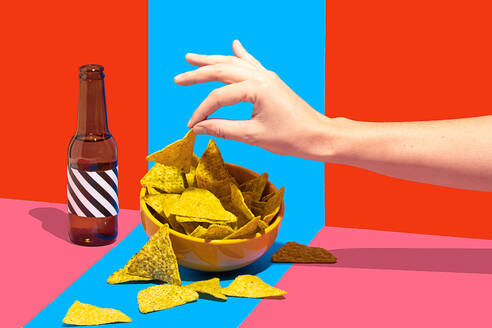 Anonymous hand reaching for nacho chips in a bowl with a bottle on a two tone red and blue background creating a dynamic and colorful setting - ADSF50026