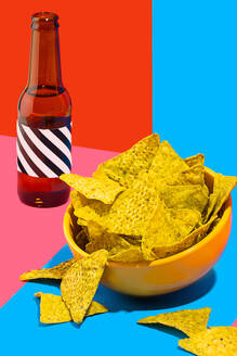 A vibrant still life image featuring a bowl of crunchy nacho chips beside a dark bottle against a split background of red and blue - ADSF50027