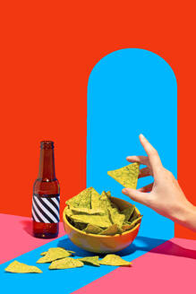 Anonymous hand reaching for nacho chips in a bowl with a bottle on a two tone red and blue background creating a dynamic and colorful setting - ADSF50028