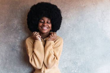 Cheerful young African American female with Afro hairstyle and with closed eyes touching sweater while standing against gray wall - ADSF50030