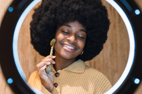 Cheerful young African American female with Afro hairstyle and closed eyes massaging face with roller facial massager during skincare routine at home illuminated by ring lamp - ADSF50031