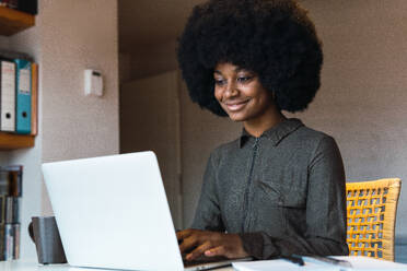 Happy African American female freelancer with stylish Afro hairstyle browsing modern netbook while working remotely at table in light room - ADSF50034