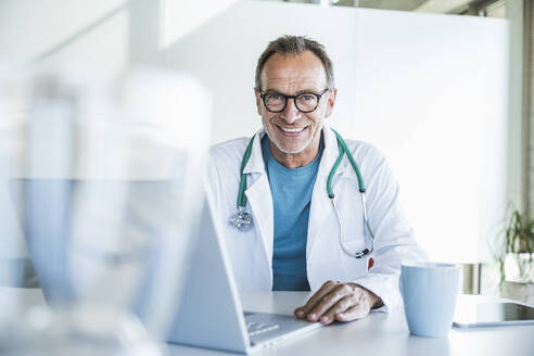 Happy senior doctor sitting with laptop at desk - UUF30610