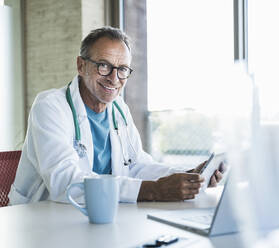 Glücklicher Arzt mit Tablet-PC und Laptop am Schreibtisch sitzend - UUF30614