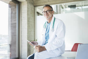 Lächelnder Arzt sitzt mit Tablet-PC am Schreibtisch - UUF30616