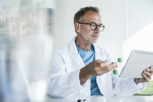 Mann im Laborkittel, der eine Molekularstruktur hält und einen Tablet-PC am Schreibtisch benutzt - UUF30620