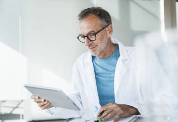 Lächelnder Arzt, der einen Tablet-PC benutzt und mit einem Notizblock am Schreibtisch sitzt - UUF30624