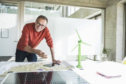 Fröhlicher Geschäftsmann gestikuliert mit Solarpanel und Tablet-PC in der Nähe eines Windradmodells auf dem Schreibtisch - UUF30641