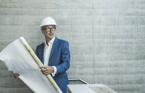 Contemplative engineer holding blueprint and standing near wall - UUF30646