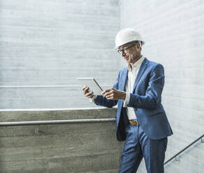 Smiling engineer wearing hardhat and using tablet PC near wall - UUF30647