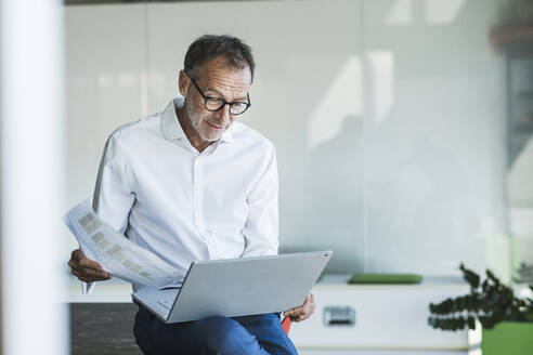 Geschäftsmann sitzt mit Diagramm und verwendet Laptop im Büro - UUF30658