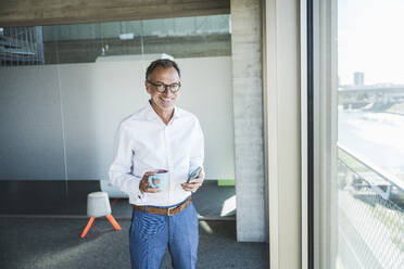 Smiling businessman standing with coffee cup and smart phone in office - UUF30668