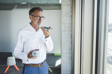 Businessman standing with coffee cup and talking on mobile phone in office - UUF30669