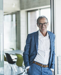 Happy businessman standing with hands in pockets at office - UUF30678