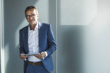 Smiling businessman standing with tablet PC in front of wall - UUF30683