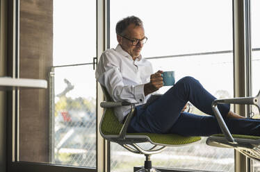 Senior businessman holding coffee cup and using tablet PC on chair - UUF30689