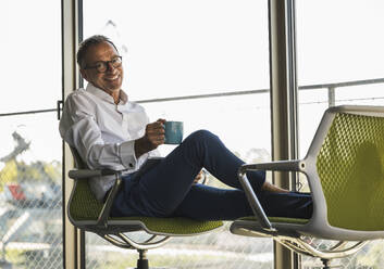Glücklicher Geschäftsmann hält eine Kaffeetasse und sitzt mit einem Tablet-PC auf einem Stuhl - UUF30690