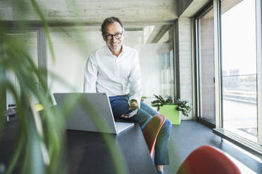 Happy businessman sitting with laptop and smart phone on desk in office - UUF30692