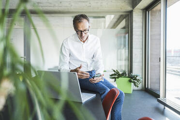 Businessman using smart phone near laptop on desk - UUF30693