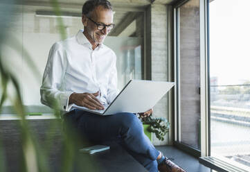 Happy businessman using laptop with smart phone on desk - UUF30696