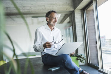 Happy businessman holding eyeglasses and sitting with laptop on desk - UUF30697