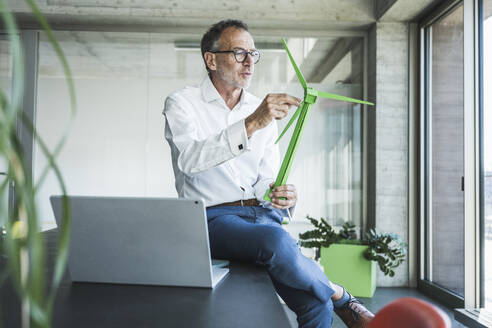 Businessman examining wind turbine model near laptop on desk - UUF30698