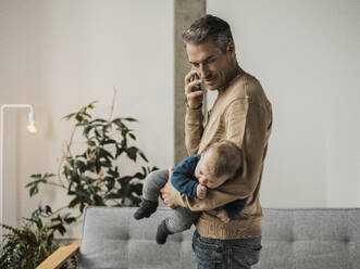 Mann hält einen kleinen Jungen und spricht mit seinem Smartphone zu Hause - MFF09512