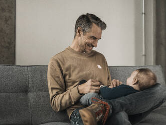 Happy father playing with baby boy on sofa at home - MFF09518