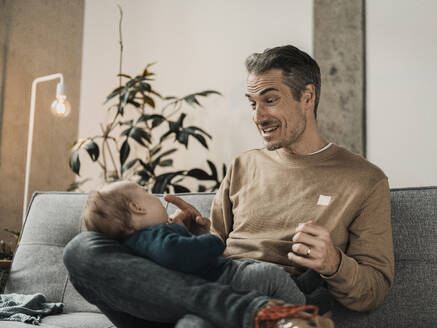 Happy father playing with baby boy in living room at home - MFF09519