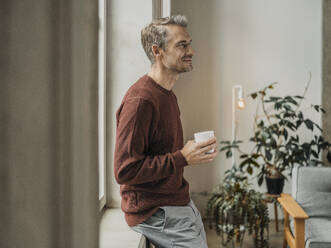 Lächelnder Mann mit Kaffeetasse, der sich zu Hause auf die Fensterbank lehnt - MFF09531