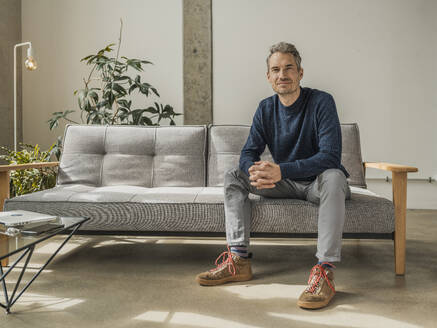 Mature man sitting on sofa with hands clasped at home - MFF09543