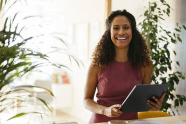 Happy businesswoman holding tablet PC near plants in office - EBSF04116