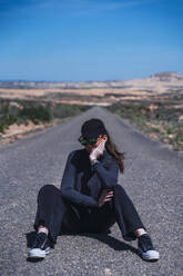 Young woman wearing sunglasses and sitting on road - RSGF00975