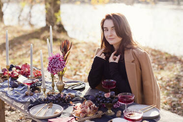 Woman wearing overcoat and sitting at table for thanksgiving celebration in forest - ONAF00699