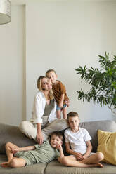 Mother with three sons sitting on sofa in living room at home - SVKF01721