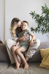 Mother kissing son sitting on sofa at home - SVKF01723