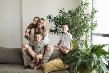 Smiling blond mother and children sitting on sofa at home - SVKF01727