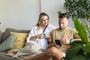 Blonde Frau mit Kaffeetasse sieht Mann an, der zu Hause sein Smartphone benutzt - SVKF01746