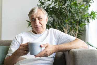 Lächelnder älterer Mann mit Kaffeetasse auf dem Sofa im Wohnzimmer zu Hause - SVKF01749