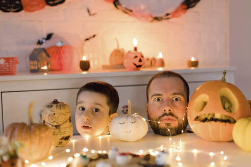 Vater und Sohn machen Gesichtsausdrücke mit dem Kopf auf dem Tisch in der Nähe von Halloween-Dekoration zu Hause - ONAF00708