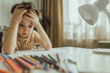 Depressiver Junge mit Papier und Bleistiften am Schreibtisch sitzend - ANAF02484
