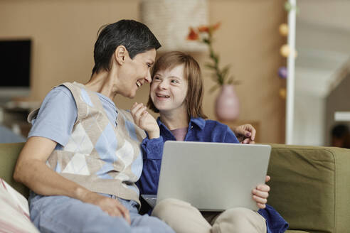 Glückliche Mutter und ihre Tochter mit Behinderung halten zu Hause einen Laptop in der Hand - KPEF00402
